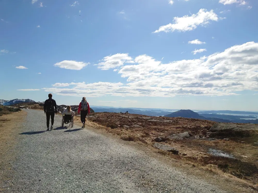 To gående og en trillende på tur på grusvei, med blå himmel og fjell i bakgrunnen