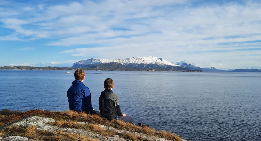To personer sitter på berget på Sandvikstien i Kristiansund med ryggen til. De har utsikt mot Tustnafjella