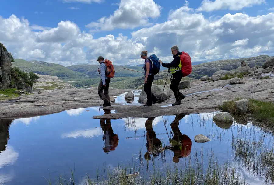 Tre personer går forbi et vann i fjell-landskap. Den bakerste personen på rekka holder i sekken til personen foran og holder en mobilitetsstokk.