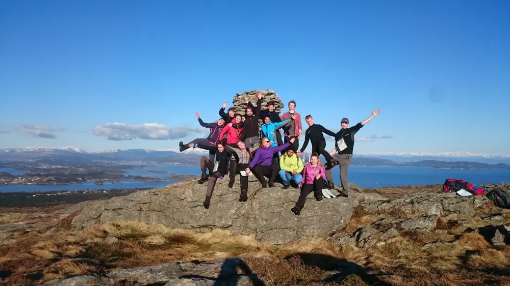 Fjorten ungdommer står foran en varde på en fjelltopp og vinker. Det er sommer, sol, og alle er glade.
