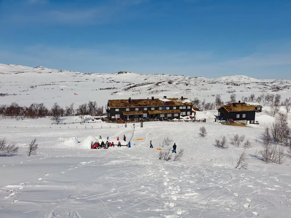 Bilde tatt fra luften ovenfra, av en langstrakt brun hytte, og to ekstra bygninger til høyre for den, på vinteren. Hele landskapet dekket av snø. En gjeng med folk på tunet foran driver og bygger en igloo, mens andre mennesker sitter på benker foran hytteveggen.