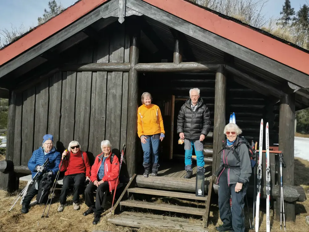 Seks eldre mennesker utenfor brun hytte.