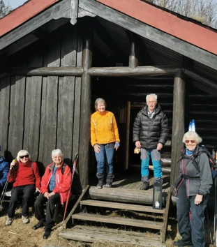 Seks eldre mennesker utenfor brun hytte.