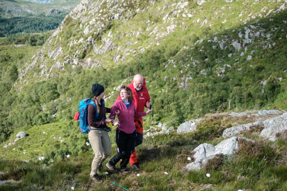 Tre personer går i fjellterreng. Damen i midten har en gul bøyle som hun holder i og blir støttet av to ledsagere. Alle personene smiler og ler.