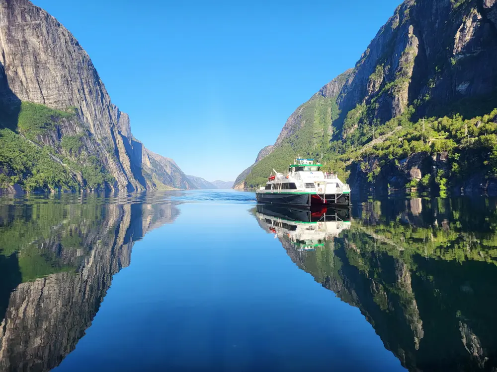 Bilde av en rutebåt som er på vei innover i Lysefjorden. Speilblank fjord med høye fjellsider.