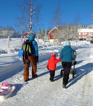 To voksne og et barn triller en barnevogn opp bakken mot en rød gårdsbygning om vinteren, med snødekte bakker rundt. Masse folk er samlet rundt hytta, og noen leker i snøen.