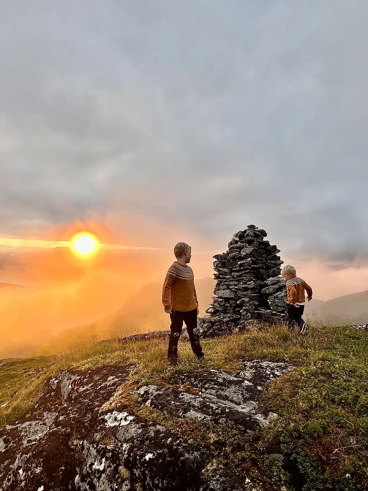 En varde på en høyde, om høsten. Lav sol i bakgrunnen gir et oransje preg. To gutter står mellom oss og varden, med ryggen til. 