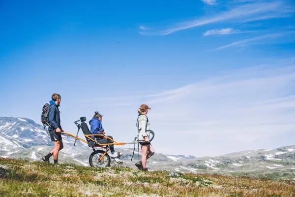 To personer på tur med en tredje i vogn for bevegelseshemmede i høyfjellet en fineværsdag.
