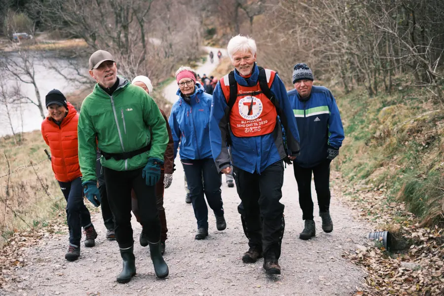 En gruppe turgåere med en turleder med rød T på brystet i front. Alle smiler og går oppover en grusti.