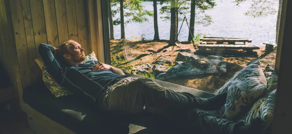En person hviler på en sofa ved et vindu som vender ut mot en skog og en innsjø. Solstråler skinner gjennom trærne og skaper en fredelig atmosfære. Personen ser avslappet ut og nyter den rolige omgivelsen inne i en hytte