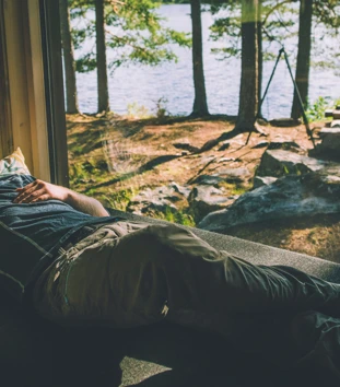 En person hviler på en sofa ved et vindu som vender ut mot en skog og en innsjø. Solstråler skinner gjennom trærne og skaper en fredelig atmosfære. Personen ser avslappet ut og nyter den rolige omgivelsen inne i en hytte