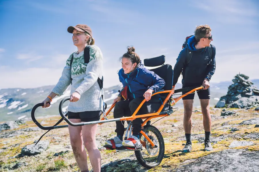 Tre personer ute på tur, der to stykker hjelper den i midten med å holde/bære en modifisert rullestol for bruk ute i naturen. 
