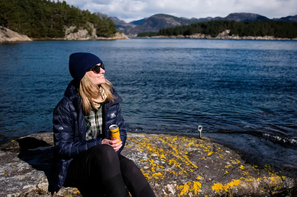 Kvinne sitter og drikker solo på berg, med sjøen bak.