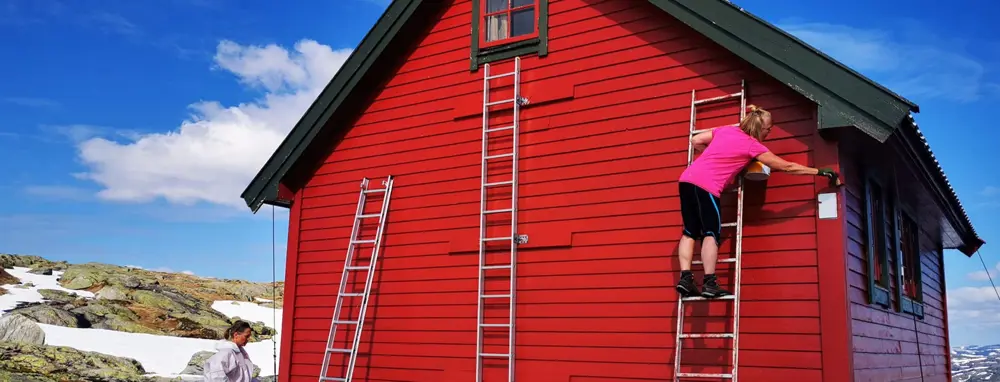 En dame i rosa genser maler en rød hytte.