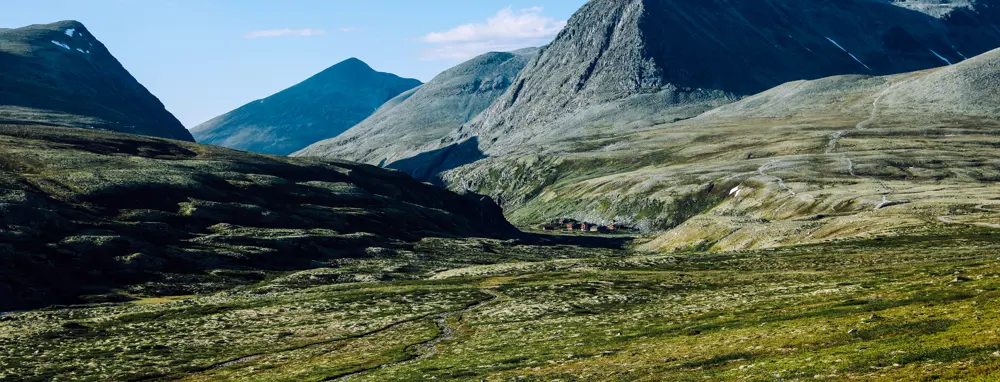 Et fjellandskap med steinete og kuperte fjell som strekker seg mot horisonten. I bunnen av dalen kan man se noen små bygninger som står samlet, omgitt av grønt landskap og lyng. Fjellene i bakgrunnen gir et majestetisk og dramatisk inntrykk, mens noen områder fortsatt har små flekker av snø.