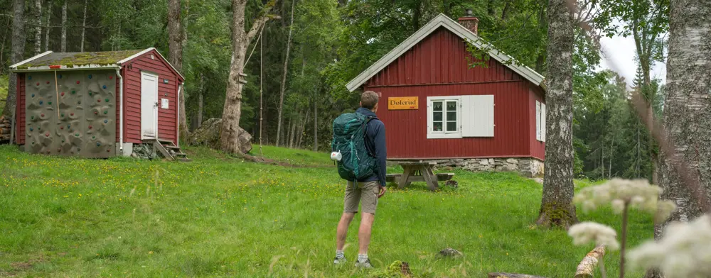 Person med sekk foran en rød hytte. 