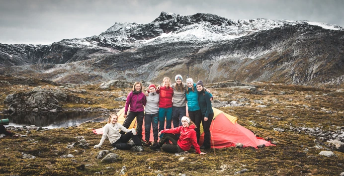 Åtte personer oppstilt foran et telt, to av dem sittende. Alle har på varme klær. Fjell i bakgrunnen.