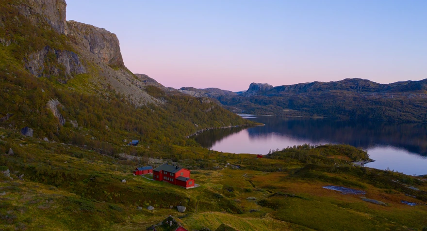 En rød hytte i solnedgang som ligger i et storslått fjellterreng med et vann i sentrum av fjellene.