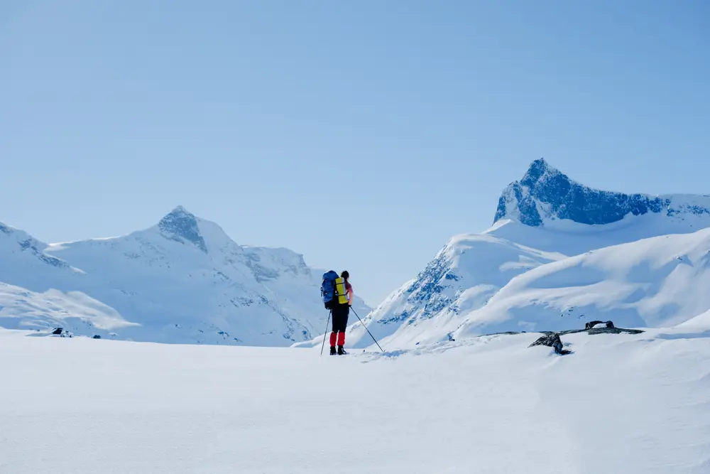 A lone skier with a large backpack looks out over a vast, snow-covered mountain range under a clear blue sky, surrounded by tall, rugged peaks.