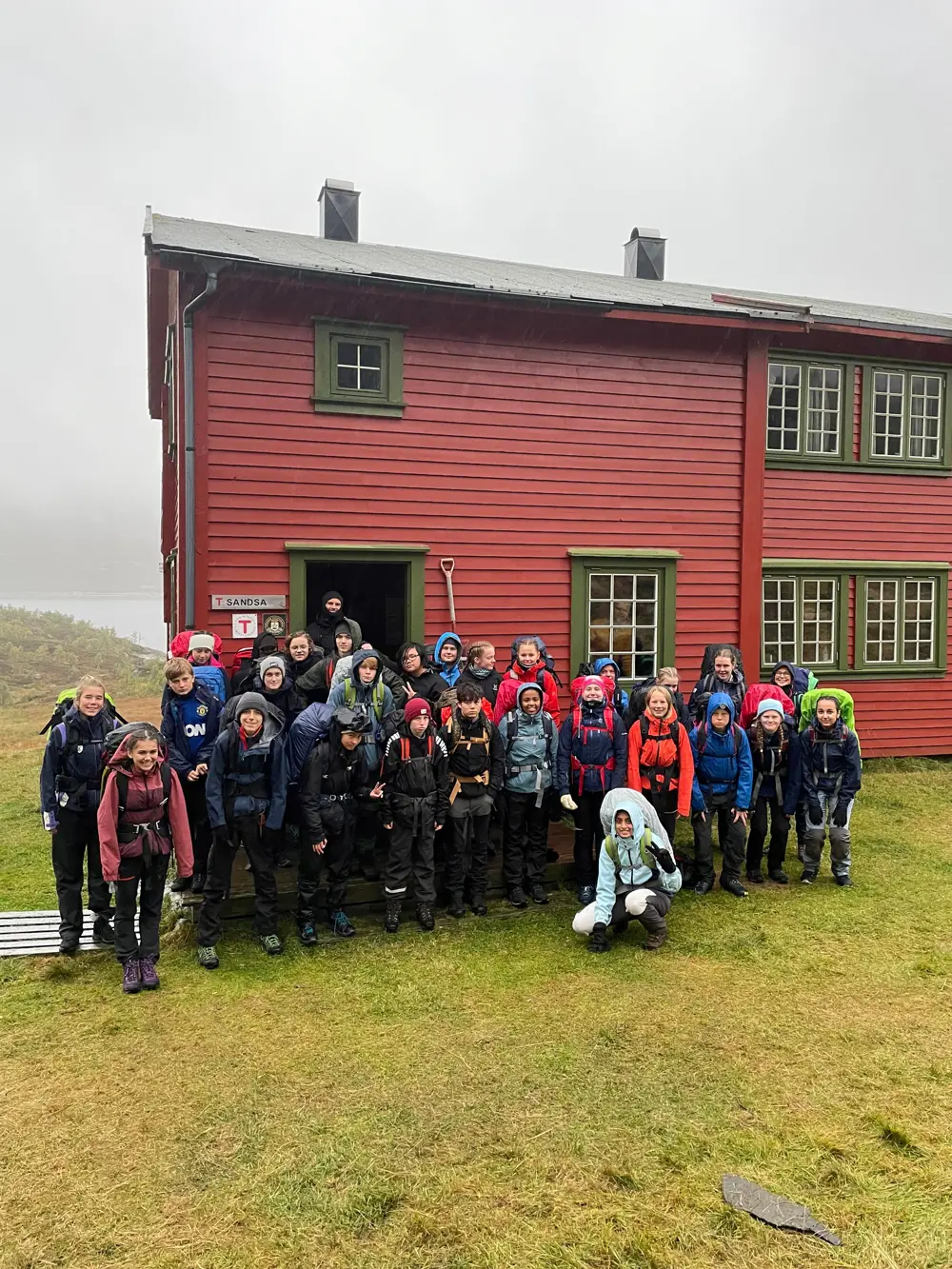 Mange ungdom med jakke og sekk  i står foran en rød hytte med grønne karmer. 