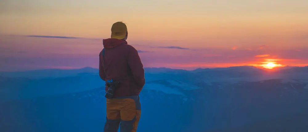 En person står med ryggen til kameraet, iført en rød jakke og gul lue, og ser ut over et fjellandskap i skumring eller solnedgang. Kameraet hans henger over skulderen, og horisonten er badet i myke lilla og rosa toner, med fjellene i bakgrunnen dekket av et blålig skjær
