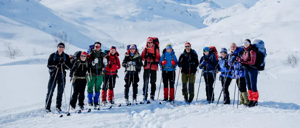 A group of smiling hikers equipped with backpacks and ski poles, dressed in winter gear, stand together on a snow-covered mountain landscape, ready for a winter adventure under clear skies