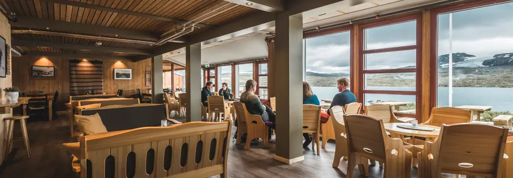 et innendørs kaféområde med store vinduer som gir utsikt over et vakkert fjellandskap. Noen mennesker sitter rundt bordene og prater, mens de ser ut mot naturen utenfor. Kaféen har treinteriør, lyse trestoler og et moderne, men koselig preg. I bakgrunnen kan man se fjell og en innsjø under en overskyet himmel