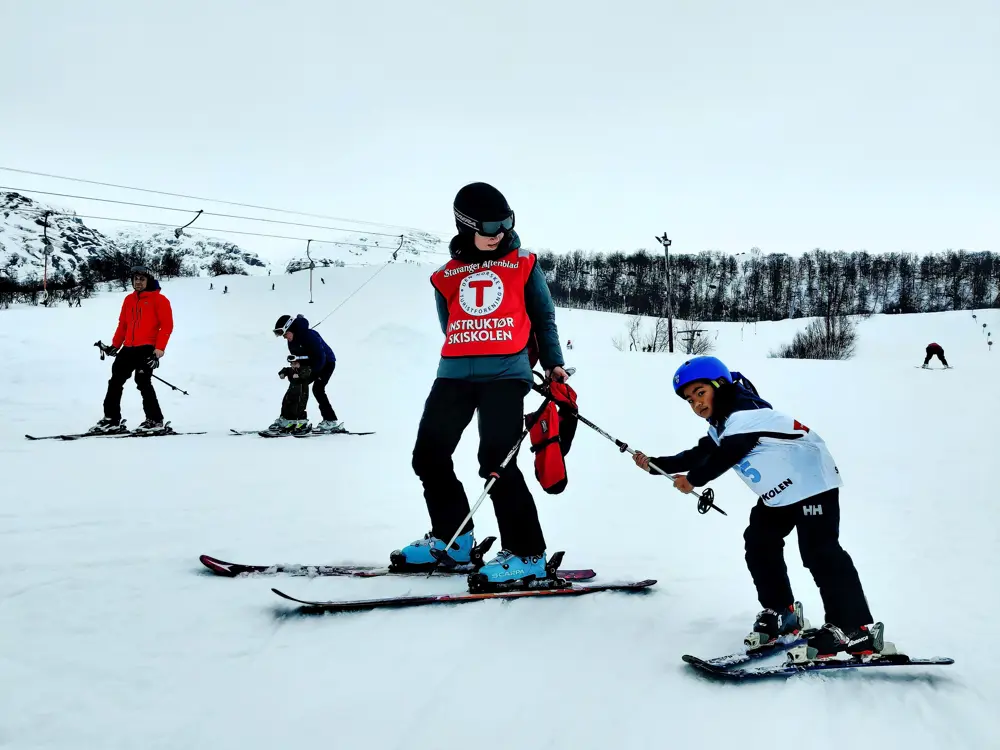 Et barn på slalomski holder seg fast i en skistav som en instruktør holder etter seg.