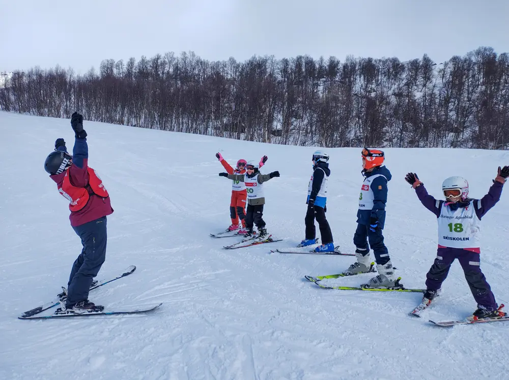 En skiinstruktør står med hendene over hodet og instruerer flere barn med slalomski på beina.