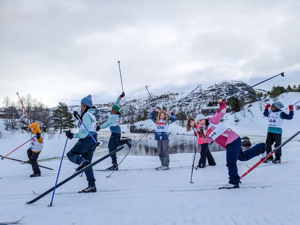 En gjeng med barn som står på langrennski vifter opp med skiene og stavene og smiler til kamera.