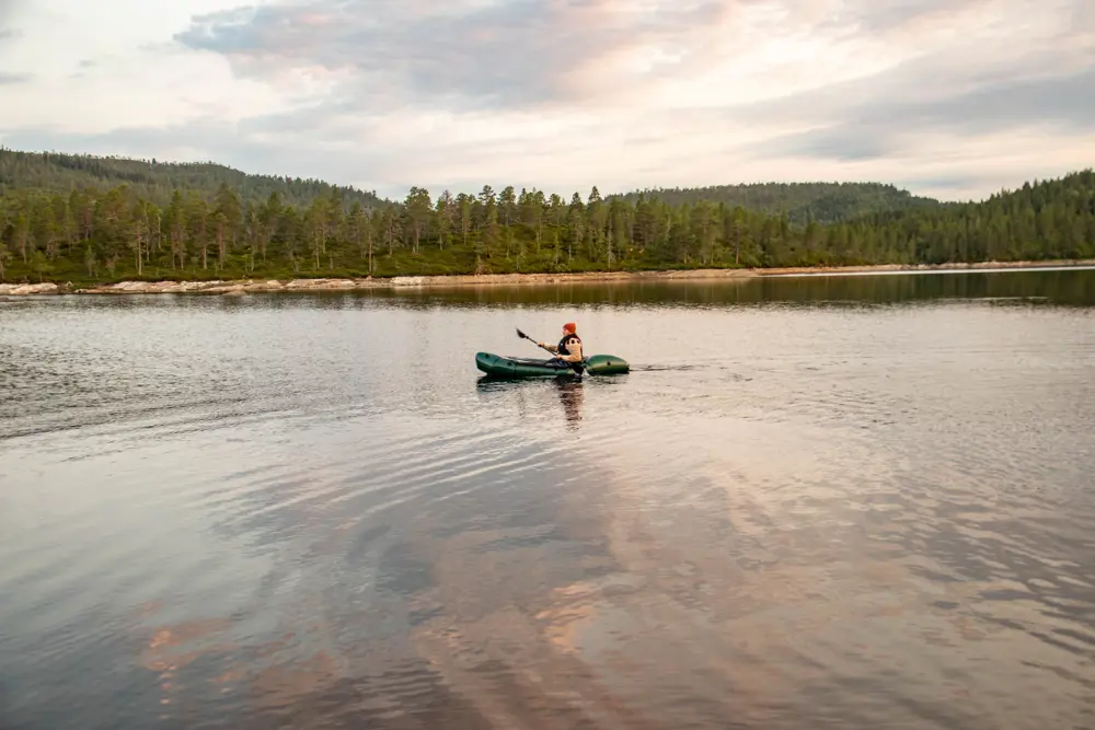Kvinne padler alene i en packraft på en innsjø, med trær langs vannbredden