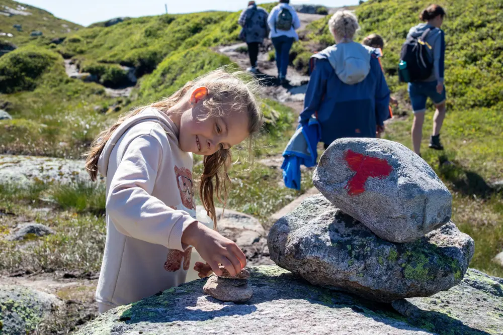 En ung jente med langt brunt hår i hestehale og hettegenser holder på med noen steiner rett ved en varde som det er malt en rød T på.