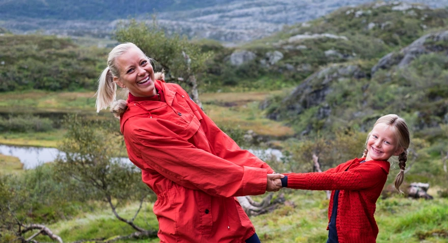 Mor og datter i rødfargede klær holder hender, lener seg tilbake og smiler mot kamera. Landskapet er grønt og på fjellet.