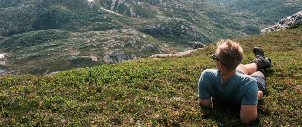 En mann som har solbriller, shorts, t-skjorte og fjellsko, ligger i gresset og ser utover fjellene