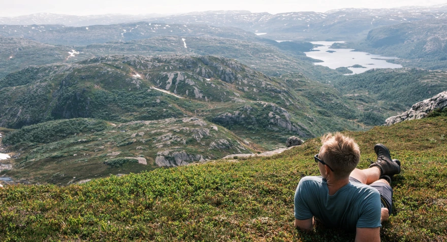 En mann som har solbriller, shorts, t-skjorte og fjellsko, ligger i gresset og ser utover fjellene