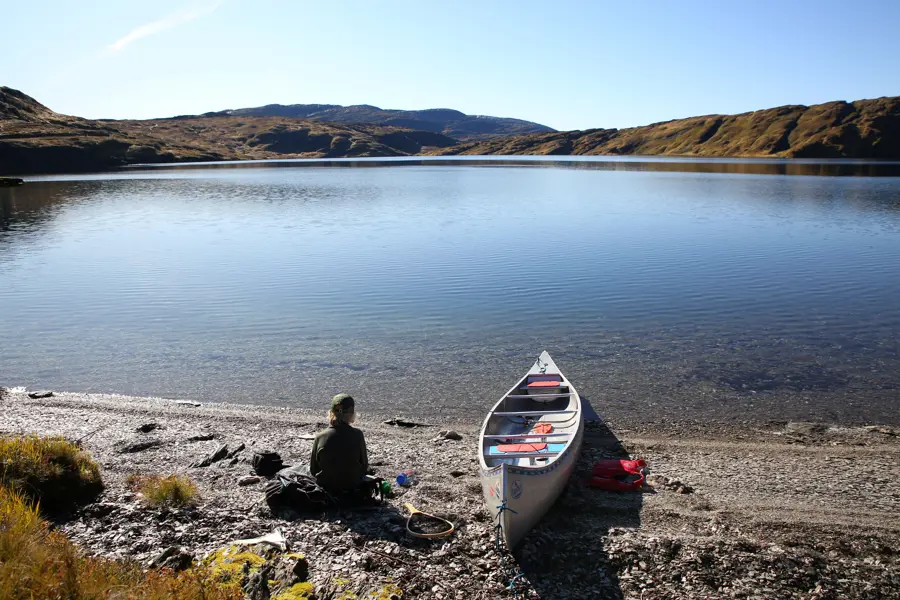 Et barn sitter på steinstrand med kano og ser utover vannet.