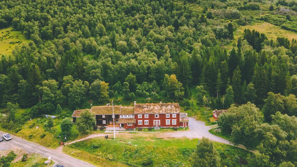 Dronebilde av en turisthytte, sett på skrå nedover ovenfra. Rød bygning med hvite kanter, omgitt av tykk skog på tre sider, og en vei som går forbi foran gressplenen. 