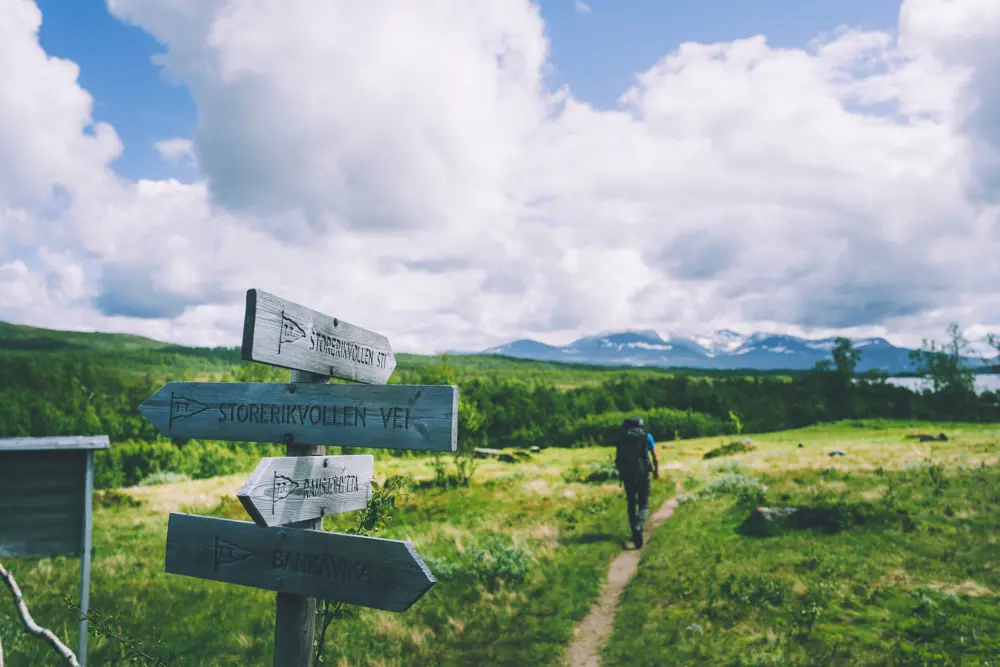 En skiltstolpe med flere skilt som peker ut retningen til forskjellige hytter. En sti fører inn i bildet, hvor en person med ryggsekk går vekk fra oss, mot skogen i det fjerne. 