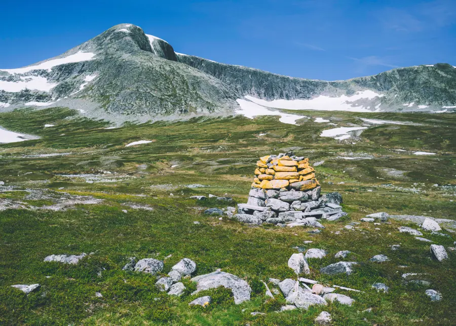 En åpen grønn slette med en varde av stablede steiner, hvor øverste halvdel av steiene har blitt malt gule. I bakgrunnen er et lite fjell. 