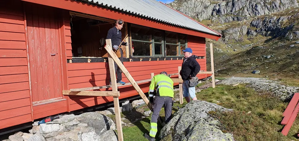 Tre menn står utenfor en hytte og bygger med planker foran en rød hytte. Det mangler et vindu i hytten. 