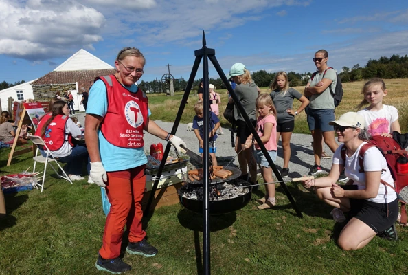 En kvinne med rød T-vest står ved en bålpanne og smiler til kamera. Mange folk rundt henne som er på et arrangement og skal grille pølser på bålpanna.