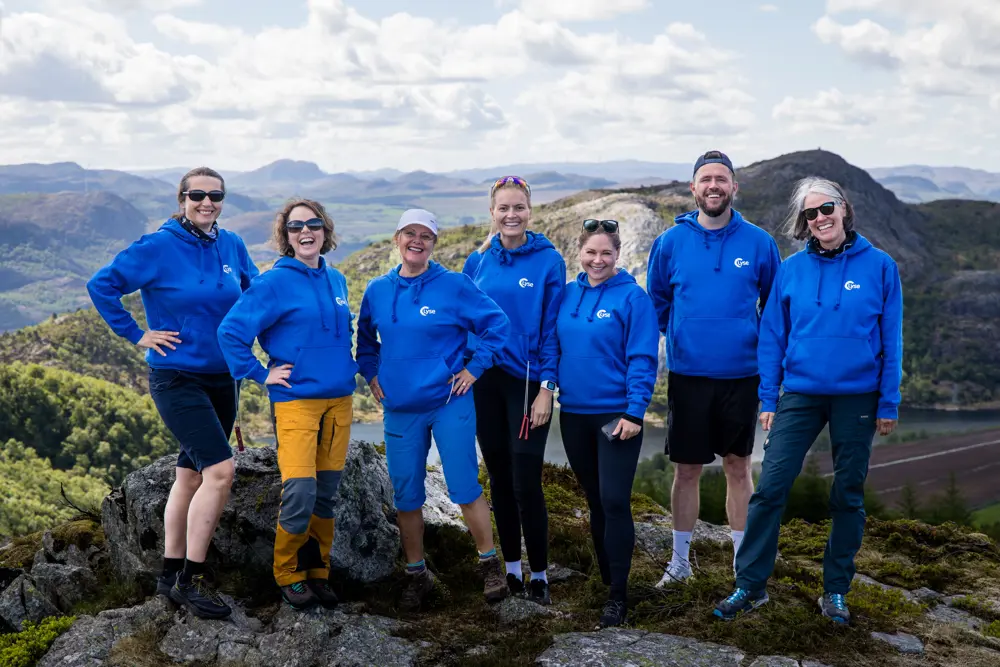 Syv glade mennesker i blå jakker smiler på toppen av et fjell