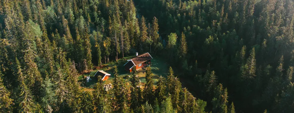 Luftfoto av en liten, rød trehytte med et hvitt skorsteinsrør, omgitt av tett skog i Nordmarka.