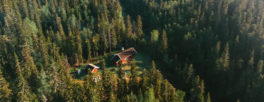 Luftfoto av en liten, rød trehytte med et hvitt skorsteinsrør, omgitt av tett skog i Nordmarka.