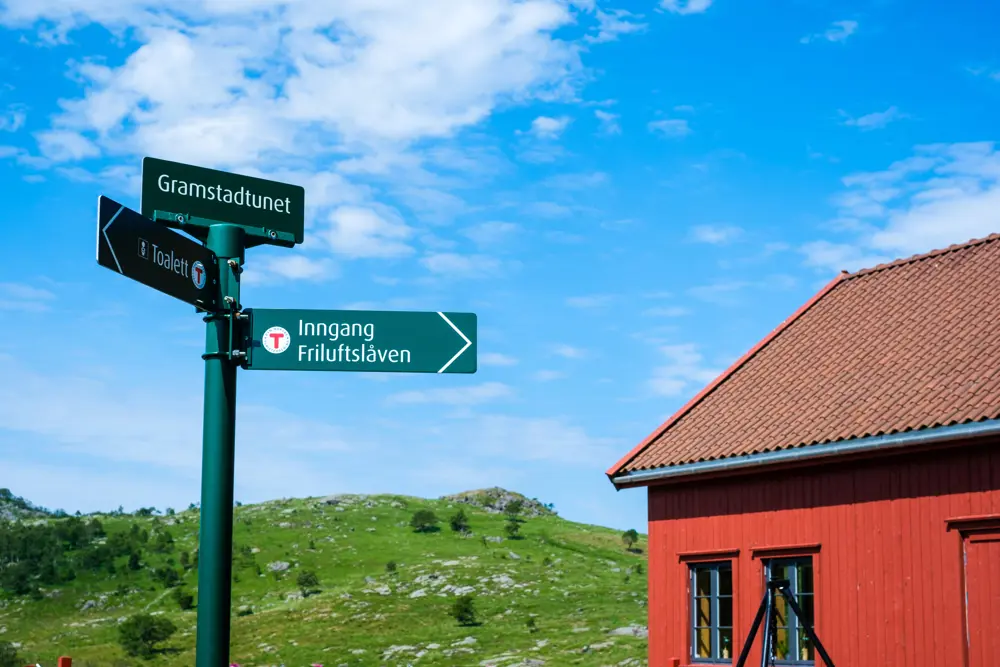 Grønne turskilt med pil til Gramstadtunet Friluftslåve. Rød låve i bakgrunnen av bilde.