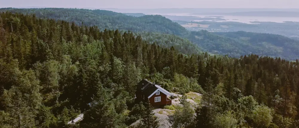 Rød hytte med hvite vinduer omkranset av tett skog og åser.