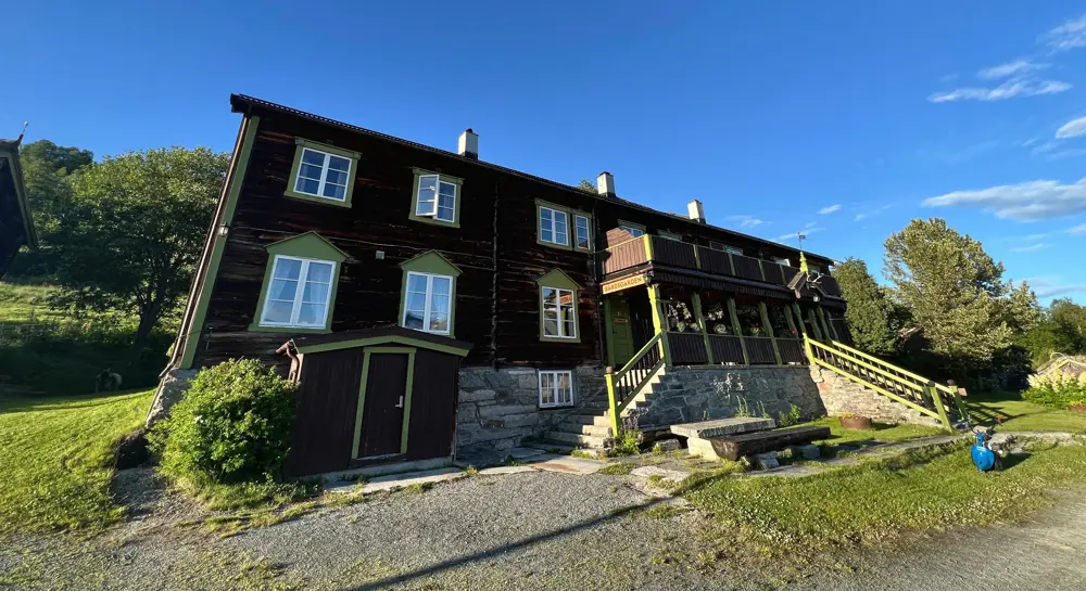 En to-etasjes brun hytte med stor veranda foran, sett fra froskeperspektiv. Skyfritt sommervær.