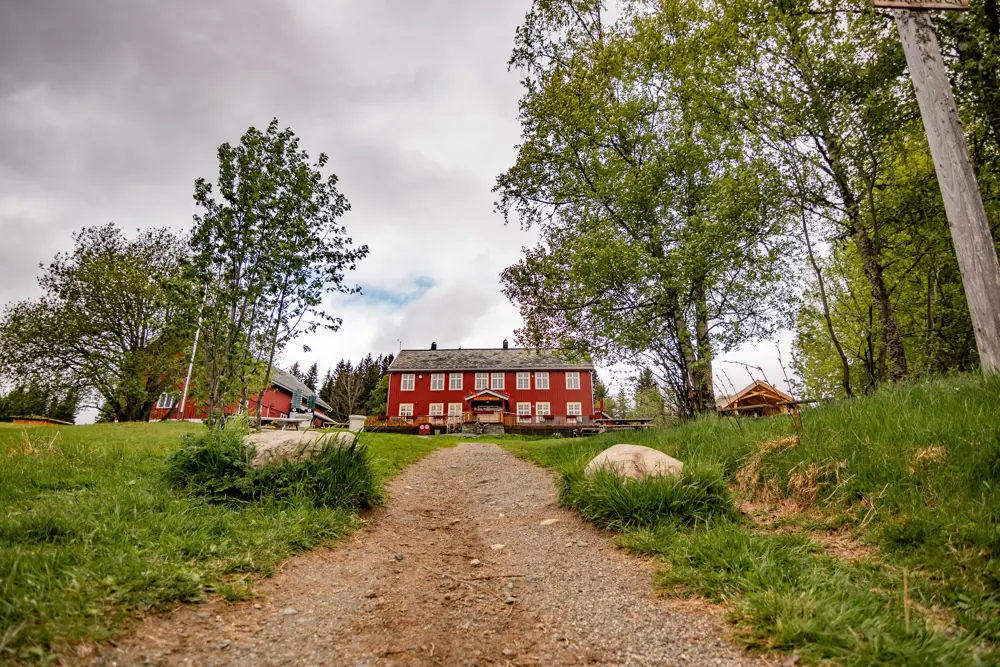 En rød stue med hvite lister og kanter er midt i bildet, og en gjørmete vei leder rett fra synspunktet og opp en bakke mot bygningen. Noen løvtrær står rundt omkring, og bakken er gressdekket. Overskyet vær. 