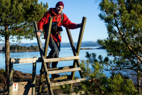 Mann i rød anorakk klatrer over gjerdeklyver med fjord i bakgrunnen. 