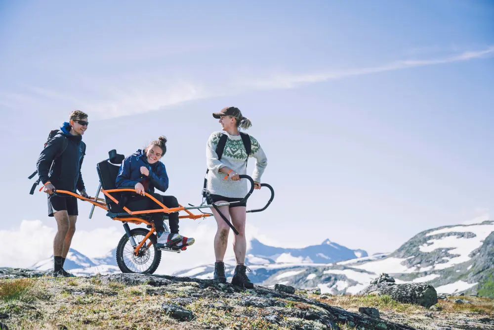 tre mennesker på tur på fjellet - to som holder turvogn og en som sitter oppi.